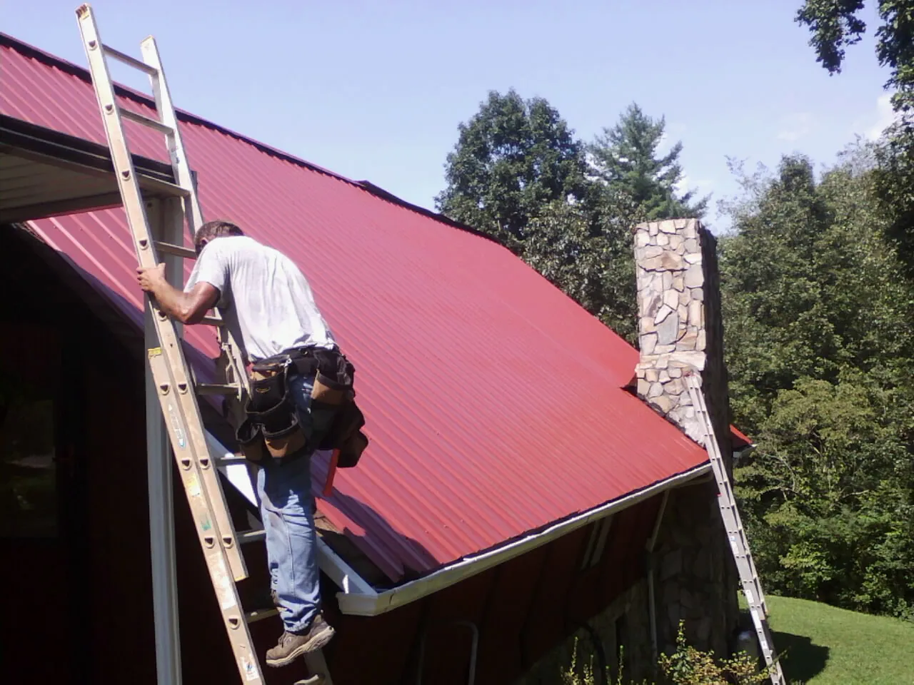 Roofer on a ladder beside a red metal roof and stone chimney — Soffit & Fascia Repair by Roofing Brothers in Dietrich